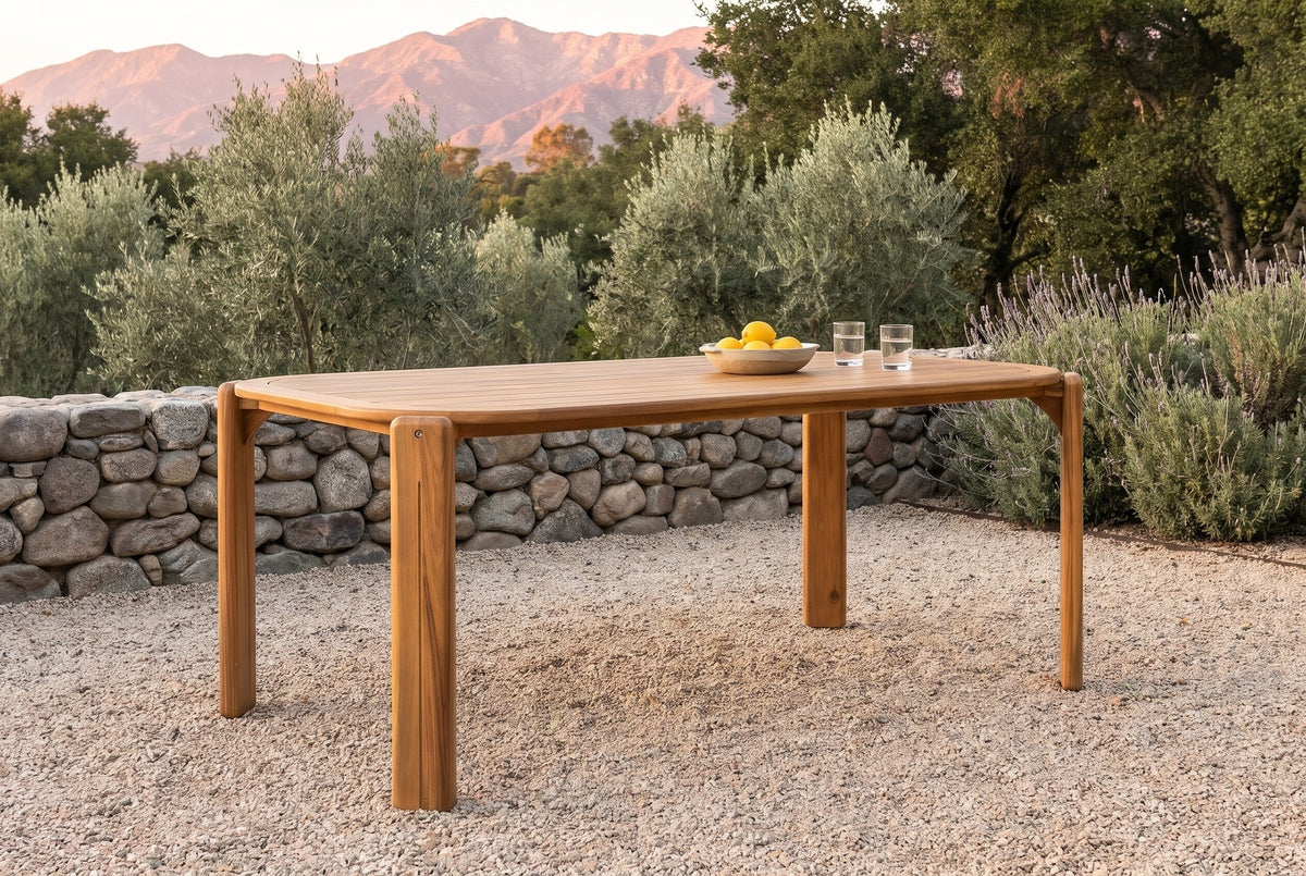 Mazara acacia outdoor dining table on pale gravel in an Ojai garden at golden hour, a dry-stacked river-stone wall, olive trees, lavender and the mountains behind (chairs not included)