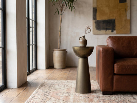 Melpignano Antique Gold Iron Pedestal Side Table beside a leather sofa in an industrial loft