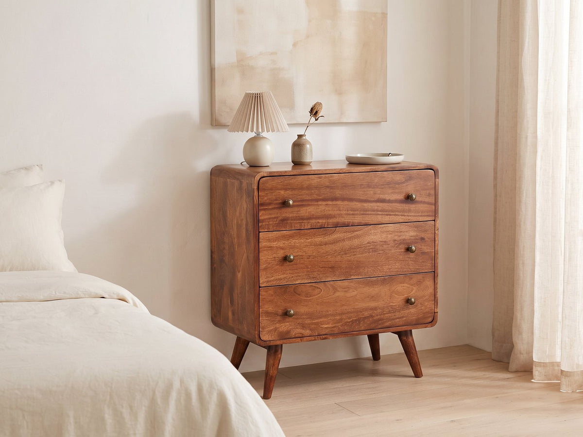 Mira 28-Inch Honey Caramel Solid Mango 3-Drawer Chest against a plaster wall under an abstract canvas in a bright bedroom