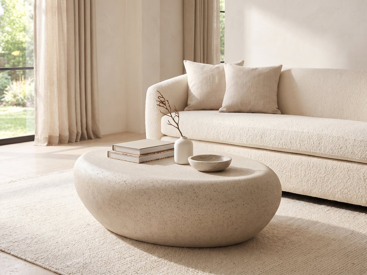Modica Cream Fiberglass Pebble Coffee Table styled with books, a vase and a stone bowl in front of a cream sofa in a bright plaster room