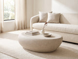 Modica Cream Fiberglass Pebble Coffee Table seen from the side, styled with books, a vase and a stone bowl in front of a cream sofa in a bright plaster room