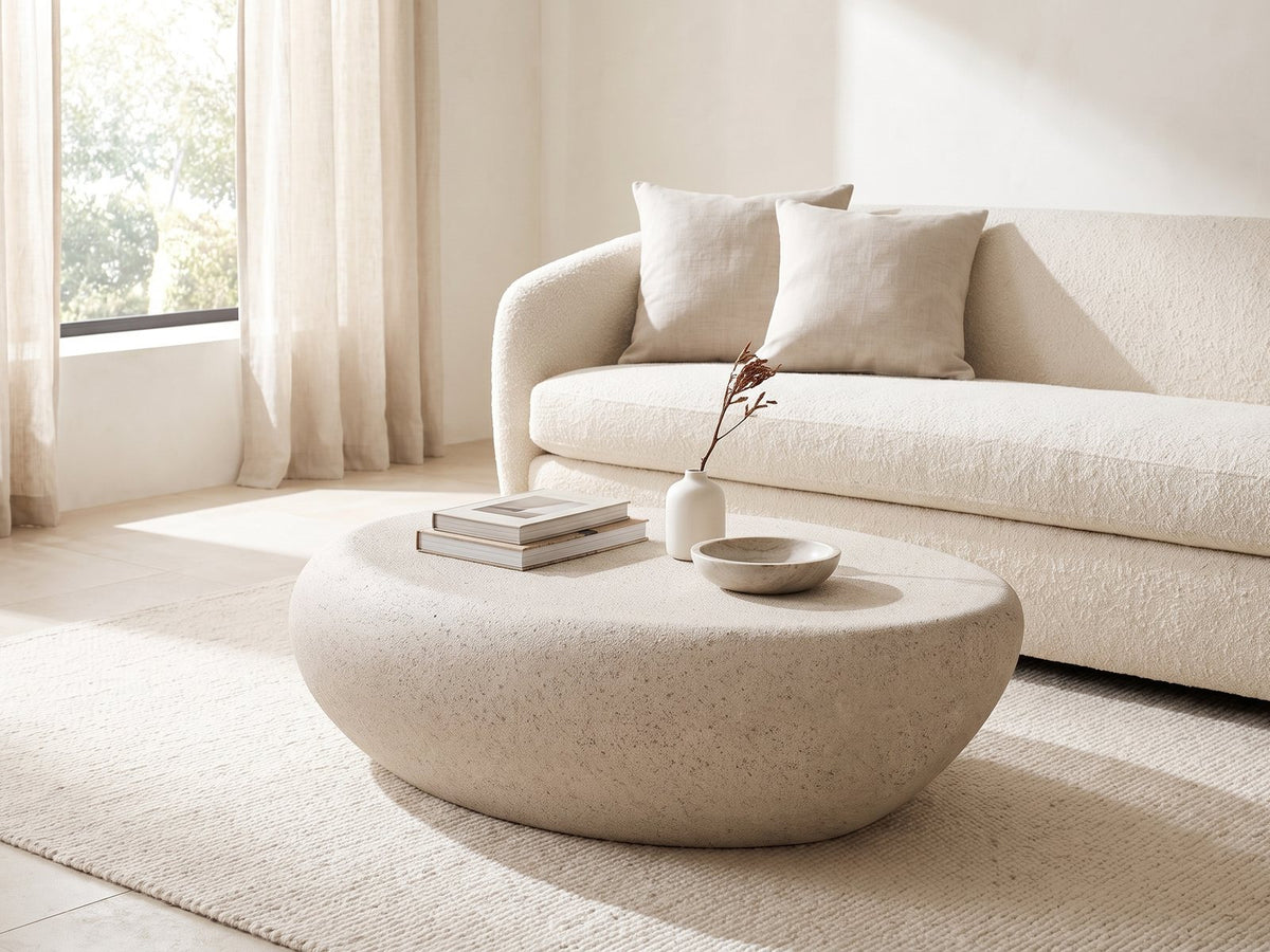 Modica Cream Fiberglass Pebble Coffee Table seen from the side, styled with books, a vase and a stone bowl in front of a cream sofa in a bright plaster room