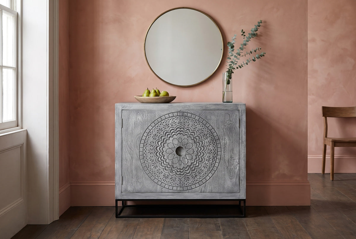 Molfetta carved grey accent cabinet against a clay-pink lime-wash wall in a Charleston dining room with a round brass mirror above it
