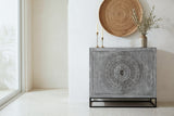 Molfetta carved grey accent cabinet with a round carved medallion on both doors, against a warm-white plaster wall in a Santa Barbara dining room with a terrazzo floor