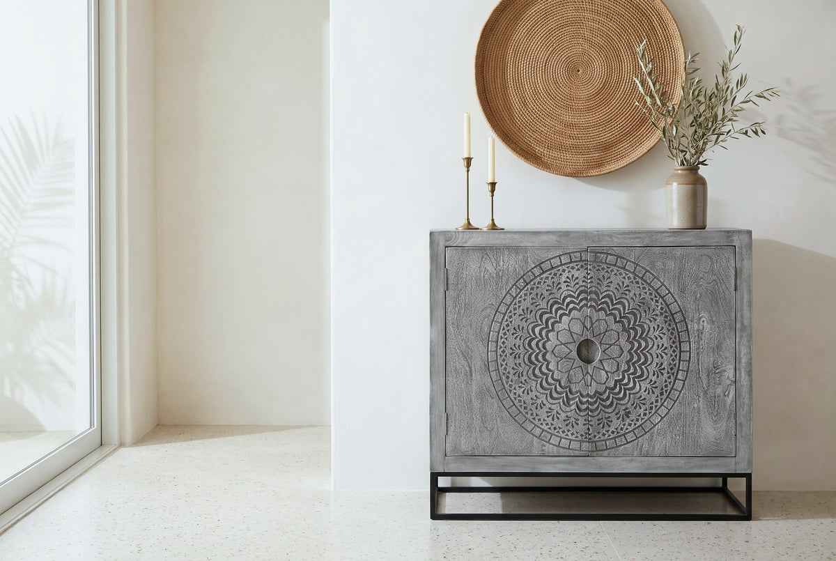 Molfetta carved grey accent cabinet with a round carved medallion on both doors, against a warm-white plaster wall in a Santa Barbara dining room with a terrazzo floor