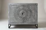 The Molfetta cabinet in sandblasted grey mango wood, front view on a light studio backdrop: two doors with one round medallion across them, on a black iron stand