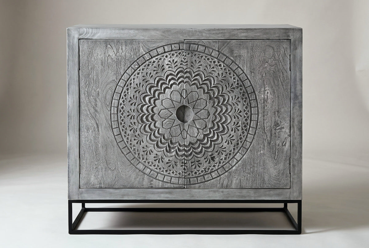 The Molfetta cabinet in sandblasted grey mango wood, front view on a light studio backdrop: two doors with one round medallion across them, on a black iron stand