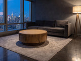 Mondovi 36-Inch Round Carved Limed Ash Drum Coffee Table on a wool rug in front of a velvet sofa in a city penthouse at blue hour
