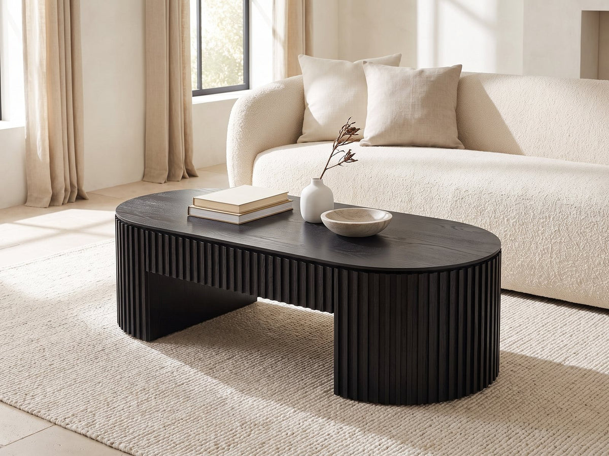 Monterosso Black Wood Veneer Fluted Oval Coffee Table styled with books, a vase and a stone bowl in front of a cream sofa in a bright plaster room