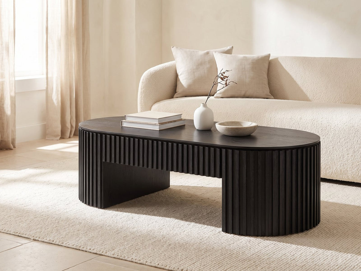 Monterosso Black Wood Veneer Fluted Oval Coffee Table seen from the side, styled with books, a vase and a stone bowl in front of a cream sofa in a bright plaster room