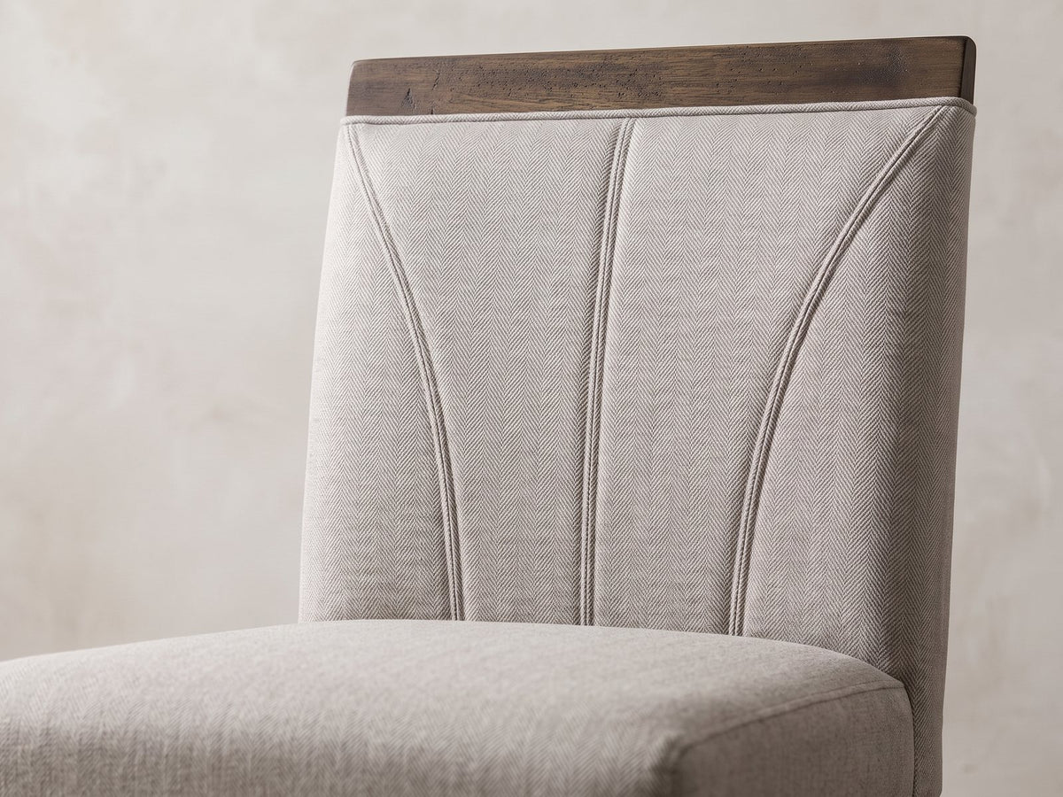 Close-up of the Mormanno 76 to 96-Inch Extending Brown Acacia Dining Set with 4 Chairs: the fan-stitched back and wood cap rail of a chair in light grey herringbone fabric