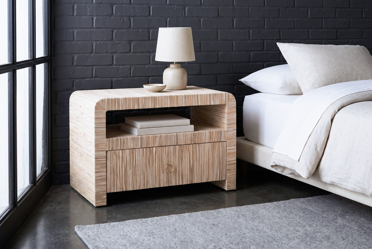 Muravera grasscloth nightstand with a ceramic lamp and two books against a charcoal-painted brick wall in a Chicago loft bedroom with a black steel window and a concrete floor