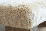 Close-up of the sheepskin on the Narni bench: long, dense natural blonde locks, pale cream at the tips over a warmer camel-beige, curving over the edge of the seat above the wood leg
