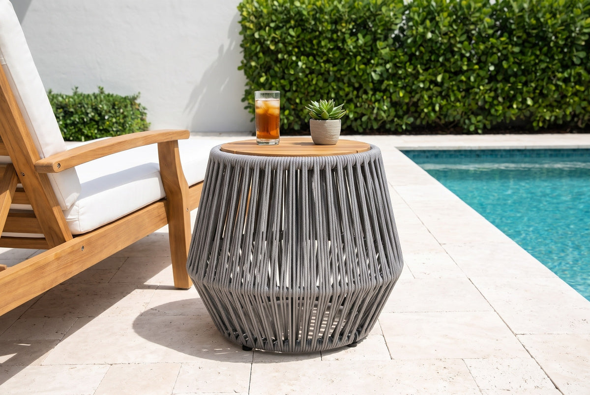 Naro grey rope and acacia side table with iced tea and a small succulent beside a teak lounge chair on a travertine pool deck in Naples, Florida, a white stucco wall and sea-grape hedge behind (chair and decor not included)