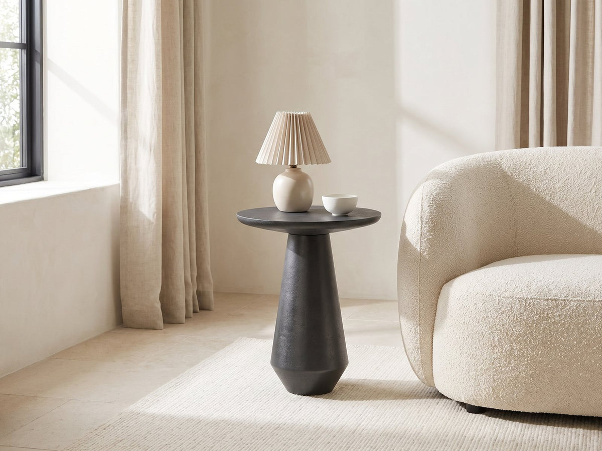 Navelli Graphite Cast Aluminum Pedestal Side Table with a lamp and a ceramic bowl beside a cream boucle armchair in a bright plaster room