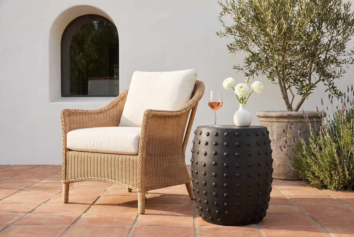 Nicolosi matte black studded side table on terracotta tiles at a Santa Barbara terrace with a white plaster wall and an olive tree, beside a woven armchair (chair, glass and vase not included)