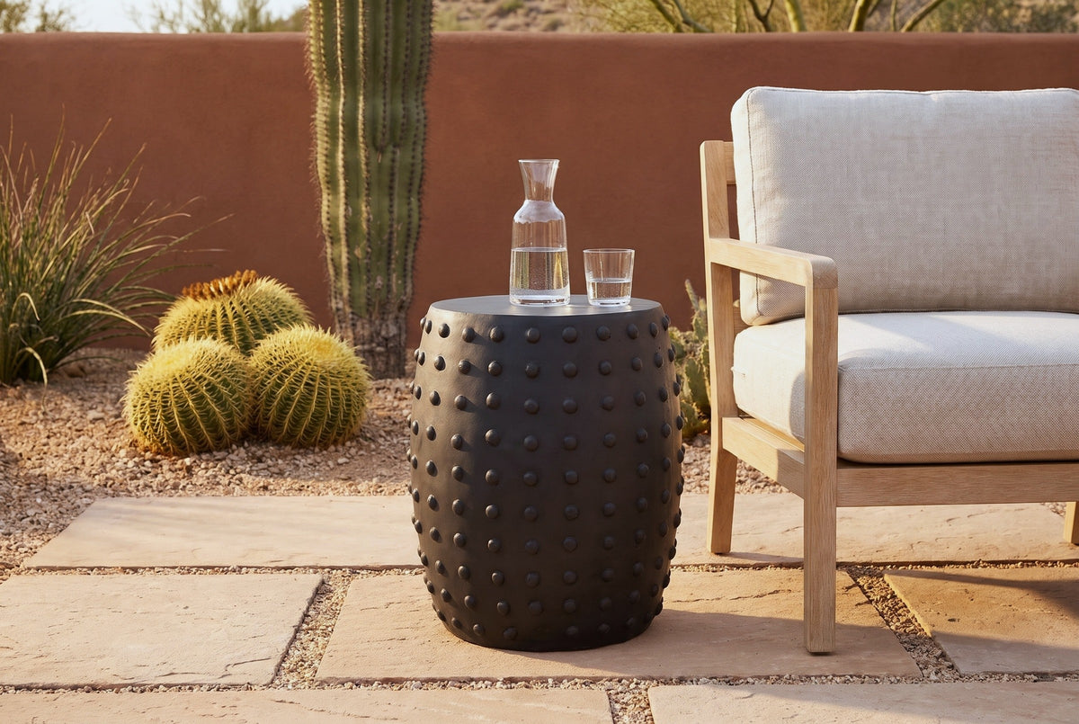 Nicolosi matte black studded side table on sandstone flagstones in a Tucson desert garden, a terracotta adobe wall, a saguaro and barrel cacti behind, beside an oak club chair (chair and carafe not included)