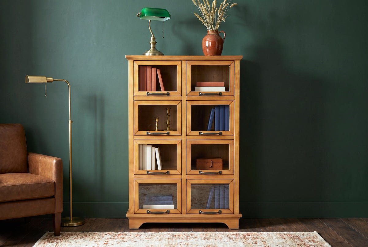 Noci pine display cabinet with a brass desk lamp and books behind its glass doors against a deep forest-green plaster wall in a Portland study with a walnut floor