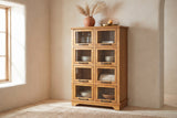 Noci pine display cabinet with pampas grass and a brass tray on top against a sand-beige plaster wall in a Santa Fe living room, with bowls, bottles and a throw behind its glass doors