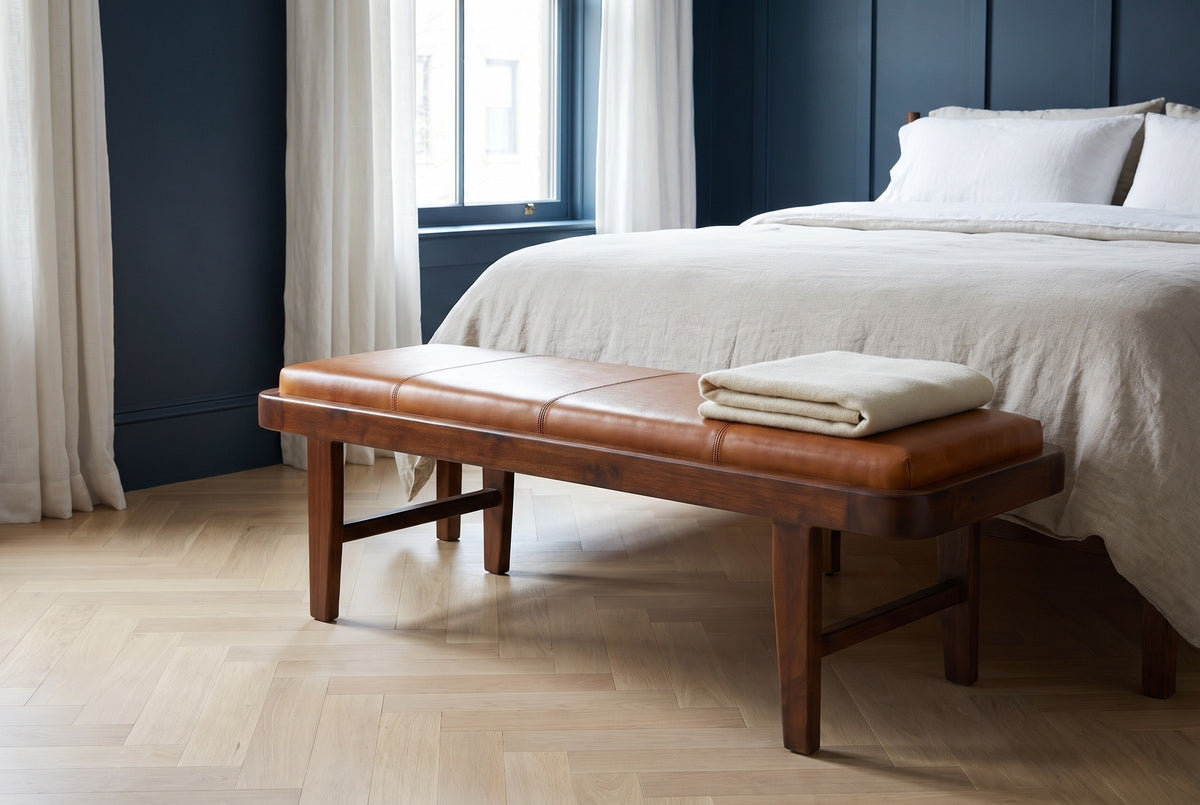 Norcia tan leather bench at the foot of a bed with white and oatmeal linen bedding in a Boston bedroom with a navy board-and-batten wall and a pale herringbone oak floor, a folded cream blanket on the bench