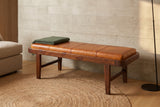 Norcia tan leather bench against a pale board-formed concrete wall in a Los Angeles living room with a round jute rug and a green wool throw