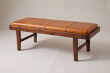The Norcia bench with its tan leather cushion in a walnut-brown wood tray, three-quarter view on a light studio backdrop