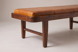 One end of the Norcia bench: the rounded end of the wood tray around the tan leather cushion, the first welted seam, two small dowel pins above the leg and the stretcher joining the two legs of that end