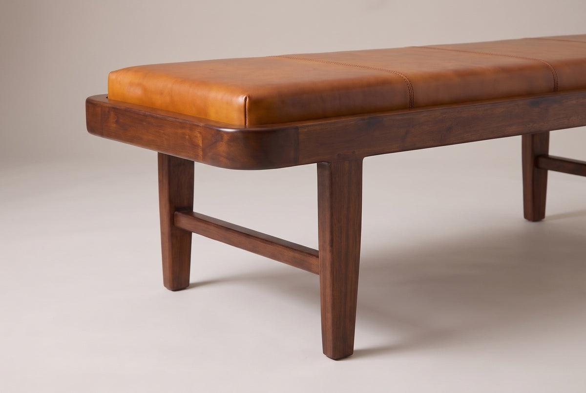 One end of the Norcia bench: the rounded end of the wood tray around the tan leather cushion, the first welted seam, two small dowel pins above the leg and the stretcher joining the two legs of that end