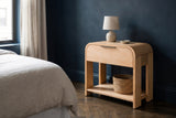 Nuoro Caramel Waterfall one-drawer nightstand with a ceramic lamp and a seagrass basket on the shelf beside a bed in a Brooklyn brownstone bedroom with a deep midnight-blue plaster wall and a herringbone floor