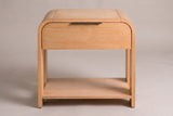 The Nuoro Caramel Waterfall nightstand, front view on a warm grey studio backdrop: a waterfall top, one drawer with a slim bronze pull, four slab legs and an open shelf