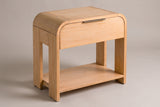 The Nuoro Caramel Waterfall nightstand with a waterfall top, one drawer with a slim bronze pull, four slab legs and an open lower shelf, three-quarter view on a warm grey studio backdrop