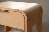 The corner of the Nuoro nightstand up close: the top rolling down over the end in a waterfall curve, the rounded corner of the drawer front and the slim bronze pull along its top edge