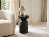 Olevano Carved Black Mango Pedestal Side Table with a lamp and a ceramic bowl beside a cream boucle armchair in a bright plaster room