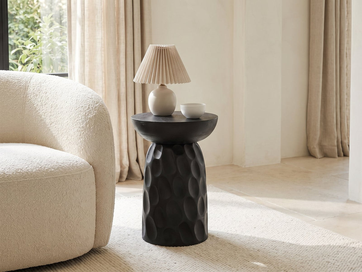 Olevano Carved Black Mango Pedestal Side Table with a lamp and a ceramic bowl beside a cream boucle armchair in a bright plaster room