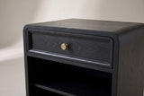 The drawer of the Oliena nightstand up close: a flat black grained panel in a raised border with an antiqued brass knob, beneath the thin top