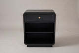 The Oliena black rounded nightstand, front view on a warm grey studio backdrop: one framed drawer with a brass knob, an open compartment with one shelf and a low plinth