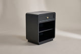 The Oliena black rounded nightstand with one framed drawer and a brass knob, an open compartment with one shelf and a low plinth, three-quarter view on a warm grey studio backdrop