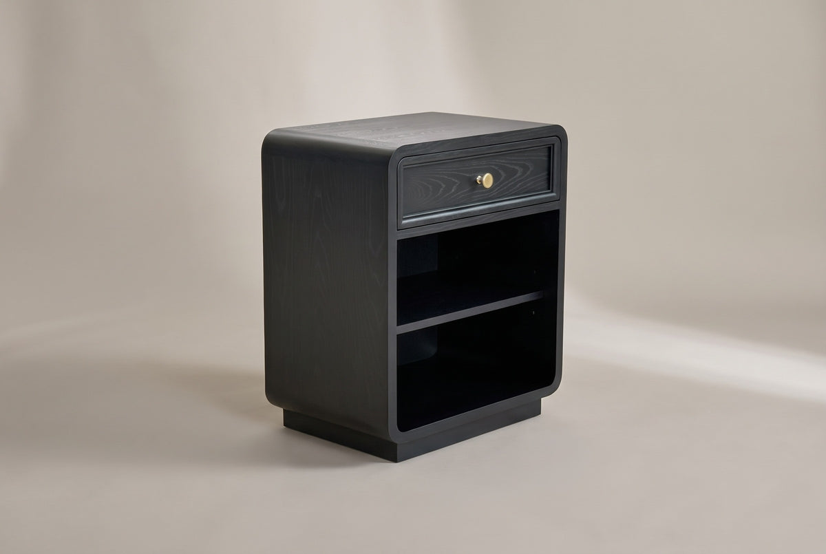 The Oliena black rounded nightstand with one framed drawer and a brass knob, an open compartment with one shelf and a low plinth, three-quarter view on a warm grey studio backdrop