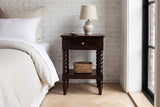 Orani dark winewood nightstand with a ceramic lamp and a seagrass basket against whitewashed brick in a Brooklyn bedroom with a herringbone oak floor and a black steel window