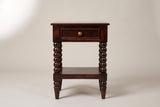 The Orani dark winewood nightstand, front view on a warm greige studio backdrop: a softly rounded top, one drawer with a brass knob, four turned bobbin legs and an open shelf