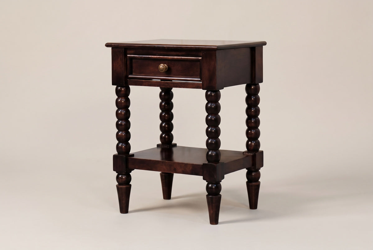 The Orani dark winewood nightstand with one drawer, a round brass knob, four turned bobbin legs and an open lower shelf, three-quarter view on a warm greige studio backdrop