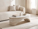 Orbetello Faux Travertine Oval Arch Coffee Table styled with books, a vase and a stone bowl in front of a cream sofa in a bright plaster room