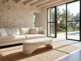 Orbetello Faux Travertine Oval Arch Coffee Table on a jute rug in front of a linen sofa in a poolside villa