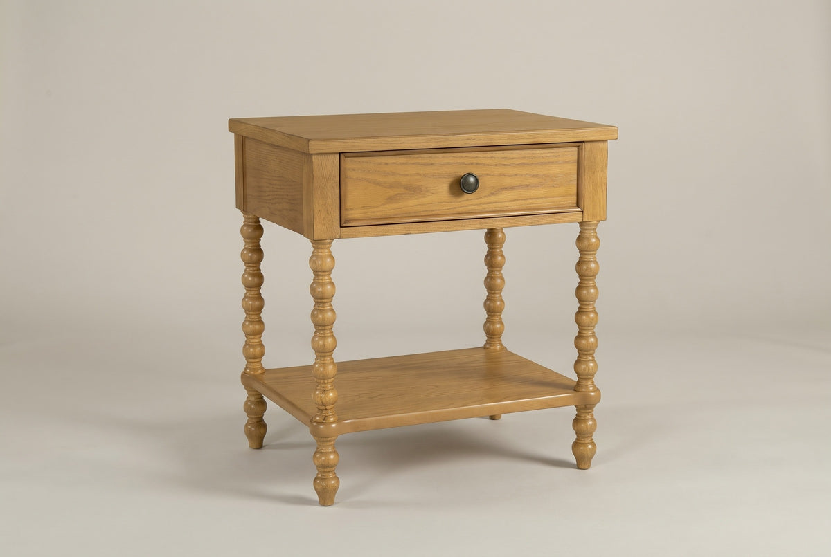 The Orgosolo Camel Oak nightstand with one drawer, a dark bronze knob, four turned bobbin legs and an open lower shelf, three-quarter view on a warm greige studio backdrop