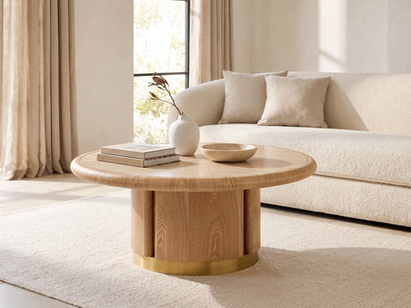 Ovada 35-Inch Round Sintered Stone and Wood Veneer Coffee Table seen from the side, styled with books, a vase and a stone bowl in front of a cream sofa in a bright plaster room