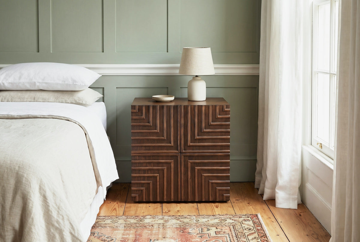 Ozieri walnut-brown ash nightstand with a lamp against a sage green panelled wall with a white picture rail in a Charleston bedroom with pine floorboards and a faded rug