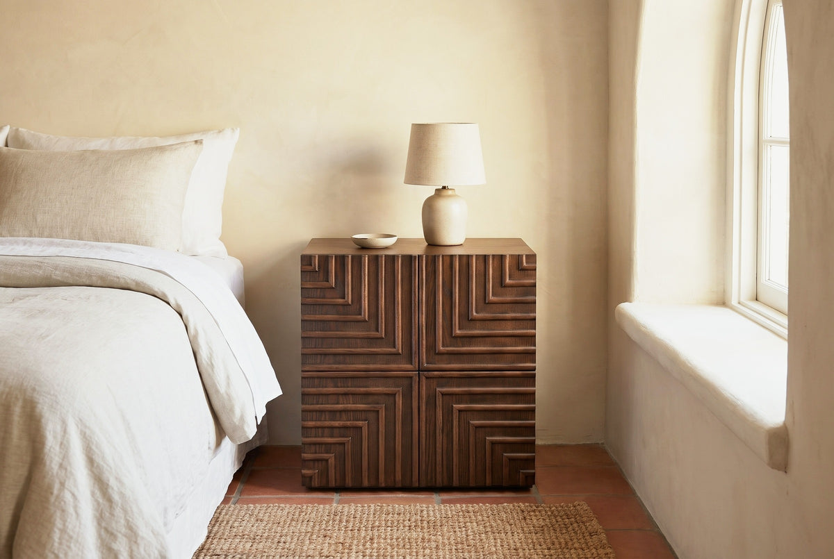 Ozieri walnut-brown ash nightstand with a lamp against a pale sand plaster wall in a Santa Barbara bedroom with terracotta tile, a jute rug and an arched window