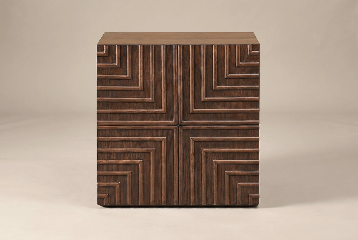 The Ozieri nightstand in walnut-brown ash, front view on a light studio backdrop: a plain top and one door carved in four mirrored quarters of nested right angles meeting in a cross