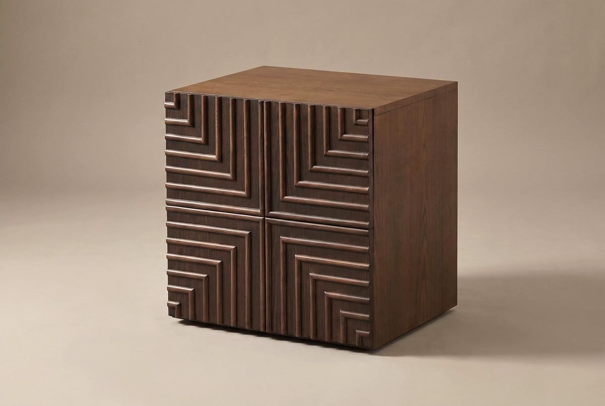 The Ozieri one-door nightstand in walnut-brown ash, three-quarter view on a light studio backdrop: a door carved in four mirrored quarters of nested right angles, a plain top and a plain side