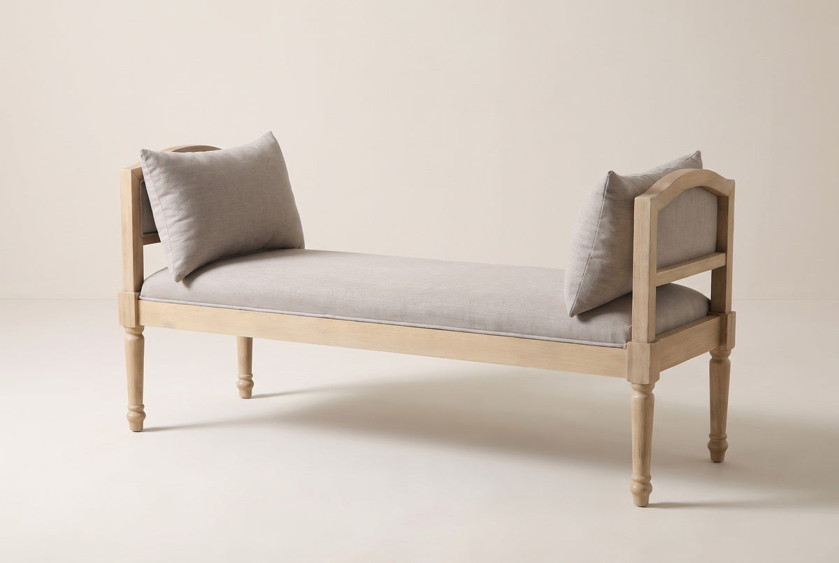 The Paciano bench in pale washed wood and greige fabric, three-quarter view on a light studio backdrop, with two pillows leaning on the arms